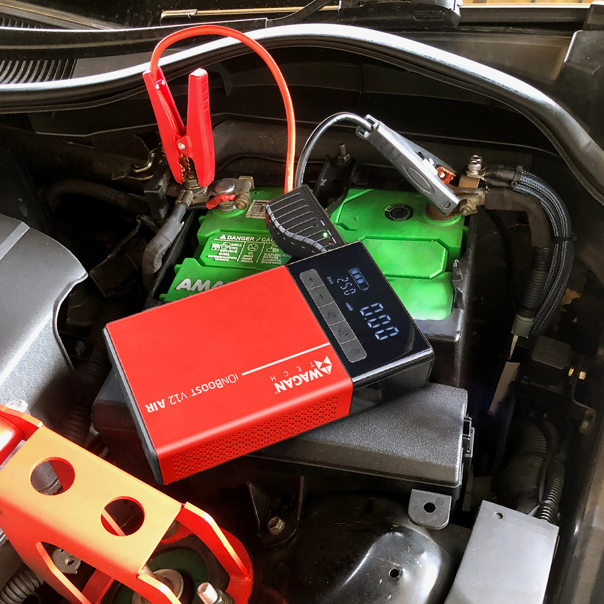 The iOnBoost™ V12 Air Compressor/Jump Starter by Wagan Tech, equipped with cables, is connected to a car battery under the hood. Serving as both a jump starter and an air inflator, the device displays numbers on its screen. The battery and engine components are visible.