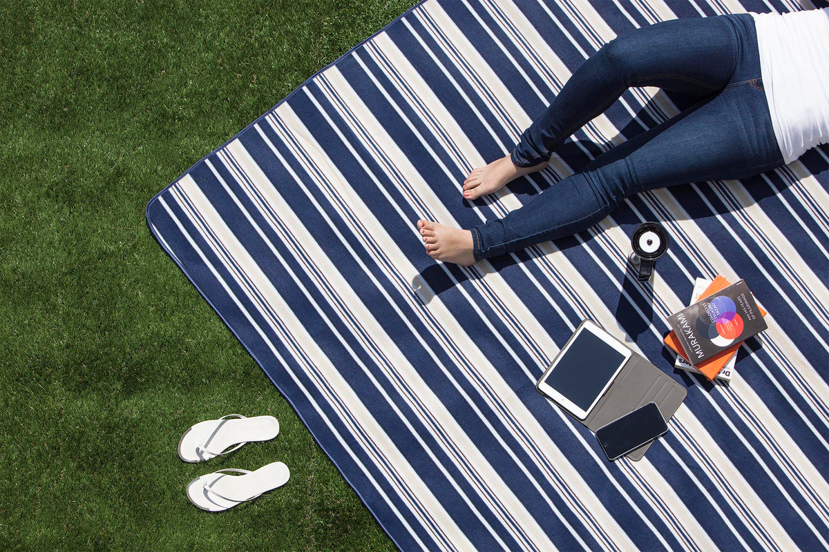 A person, visible from the waist down and dressed in jeans with bare feet, sits on an Airstream Outdoor Picnic Blanket spread across the grass. Next to them are a pair of flip-flops, a camera, an e-reader, a tablet, and a book.
