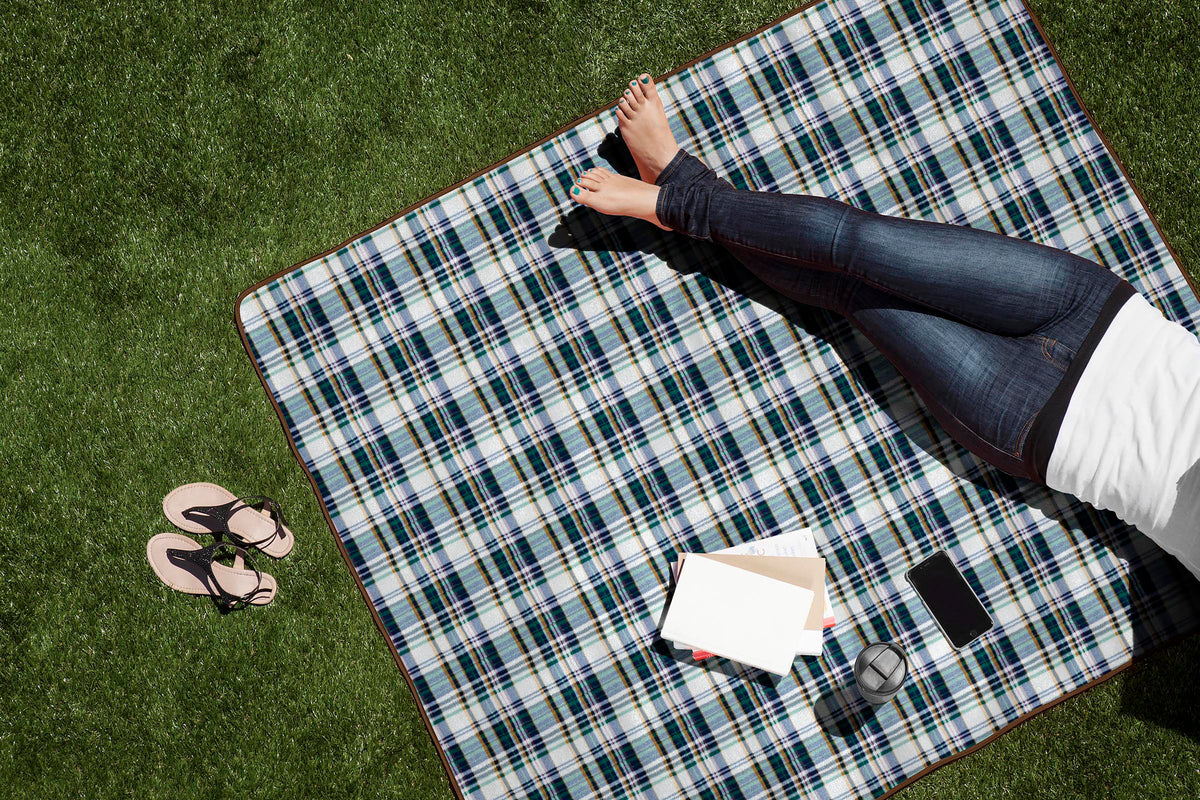 A person in jeans relaxes barefoot on an Airstream Outdoor Picnic Blanket spread out over the grass, with sandals nearby. A book, closed notebook, reusable bottle, and smartphone are placed beside them.