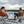 A man and a woman sit in woven camping chairs on a patterned dock, facing each other across a lake with a white portable power station between them. The scene is outdoors under a cloudy sky with trees in the distance.