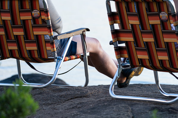 Two woven patio chairs with a striped pattern in red, orange, yellow, and brown, with a person's legs visible sitting between them outdoors near water.