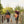 Two people walking on a wooden boardwalk with a dog, each carrying a woven orange and black folding chair over their shoulder in an outdoor park setting.