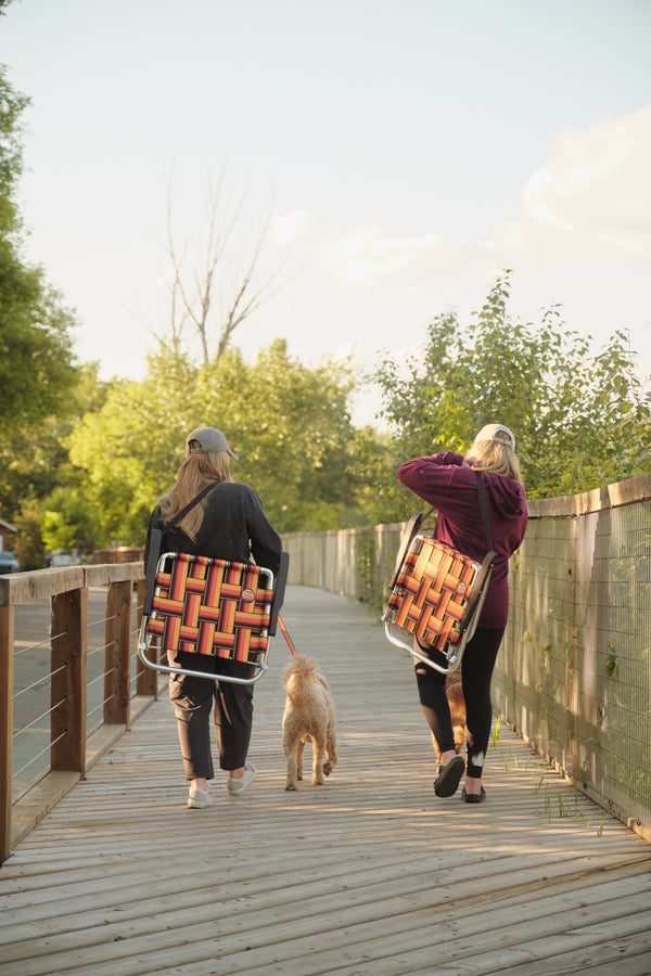 Two people walking on a wooden boardwalk with a dog, each carrying a woven orange and black folding chair over their shoulder in an outdoor park setting.