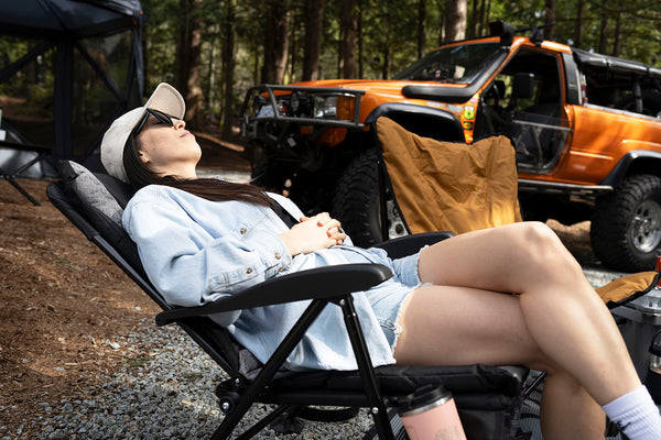 A woman relaxes in a black camping recliner chair outdoors in a wooded campsite setting, with an orange off-road truck parked in the background.