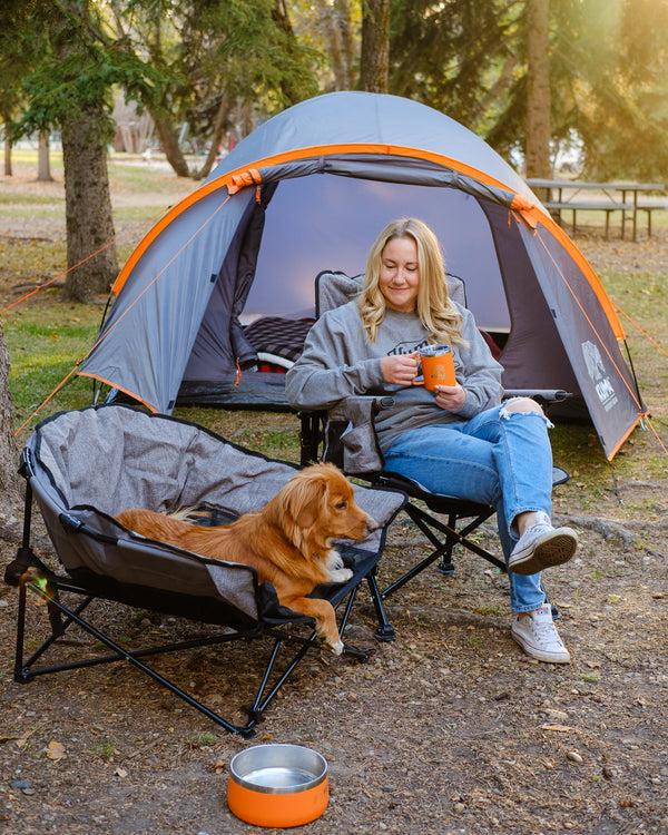 A woman sits in a camping chair in front of a gray and orange tent, holding an orange mug. A golden retriever rests in another camping chair beside her, and a metal bowl with an orange rim sits on the ground.