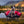 A blue heeler dog sits in a red and black camp chair placed on grass by a lake, with mountains and trees in the background.