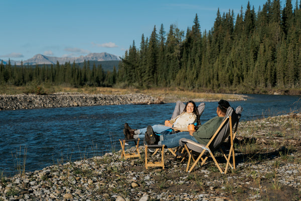 Two people relax in folding camp chairs beside a river, with a forest and mountains in the background. The chairs are light wood with dark fabric seats, set on a rocky riverbank in a natural outdoor setting.