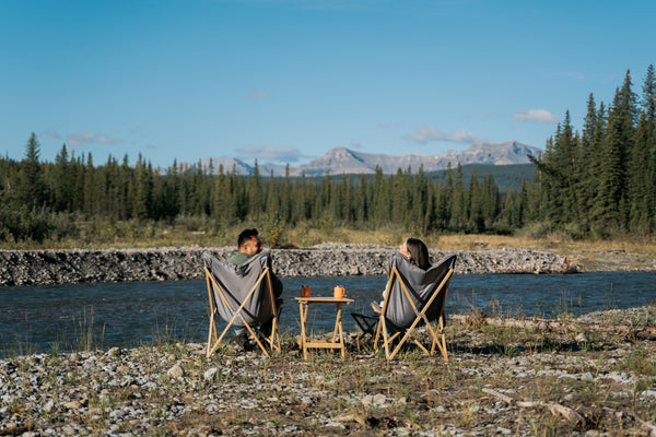 Two people sitting in folding camp chairs on a rocky riverbank, wrapped in blankets and facing a scenic mountain landscape with a lake and pine forest under a blue sky.