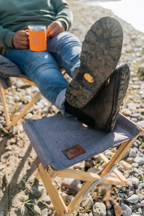 A person reclining outdoors with their legs crossed over a gray fabric camping chair, wearing dark brown rubberized boots. They are holding an orange mug, with pebbled ground and another chair visible in the background.