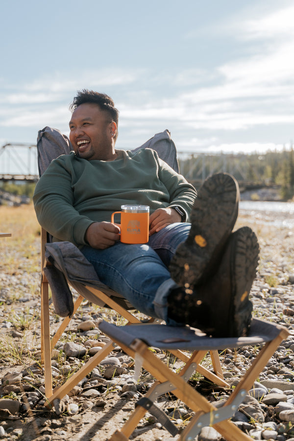 A man relaxes in a folding camp chair outdoors near a rocky riverbed, with his boots propped up. He is wearing a green long-sleeve shirt, blue jeans, and holding an orange mug.