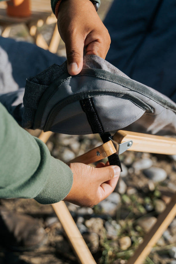 A close-up lifestyle shot of a person adjusting the strap of a gray and black backpack on a wooden folding camp chair outdoors.