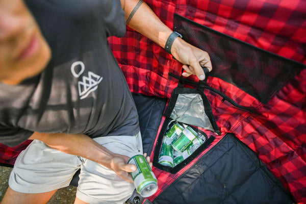 A close-up lifestyle shot of a person organizing green beverage cans inside a red and black buffalo plaid cooler bag or tent pocket.