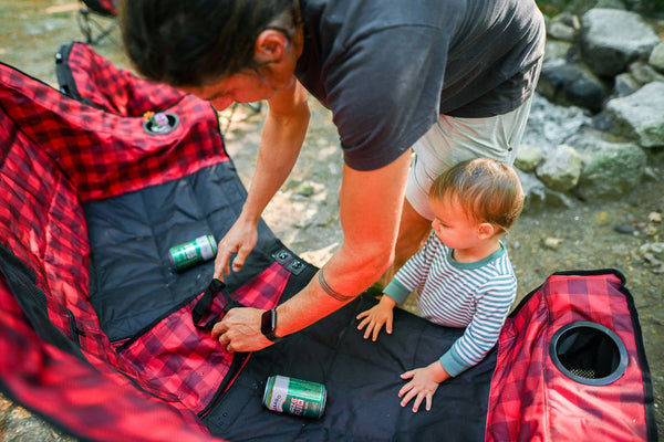 A man and a young child are setting up a red and black plaid camping blanket or mat outdoors near rocks. The man is arranging the fabric while the child watches, with two green beverage cans placed on the mat.