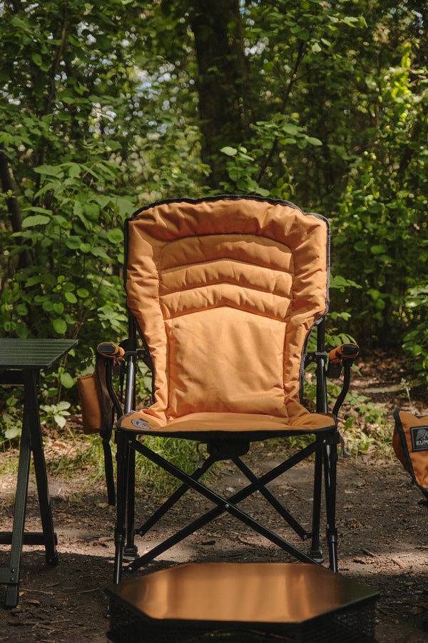 An orange padded camping chair with a black metal frame, set in a wooded outdoor environment.