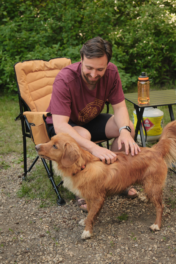 A man sitting in an outdoor camping chair pets a golden retriever dog on a gravel path with green foliage in the background.