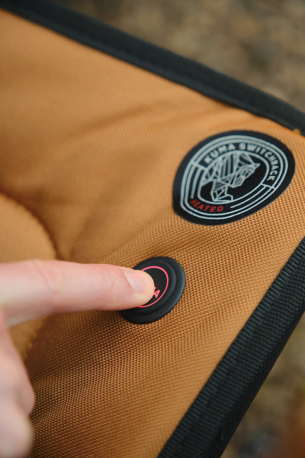 Close-up of a person's finger pressing a black power switch on a tan and black camping chair, with a circular embroidered logo patch visible on the fabric.