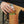 A tan canvas beverage holder pocket attached to a black folding chair, holding a green can of beer, with a person's hand reaching in.