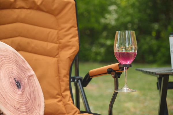 An outdoor camping chair in warm tan with a wine glass resting on its armrest, filled with pink wine, set against a blurred green garden background.