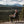 A man sits in a black camping folding chair on a rocky hillside, looking out over a vast pine forest and distant mountains under a cloudy sky.