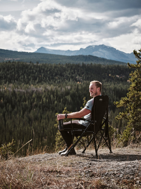 A man sits in a black camping folding chair on a rocky hillside, looking out over a vast pine forest and distant mountains under a cloudy sky.