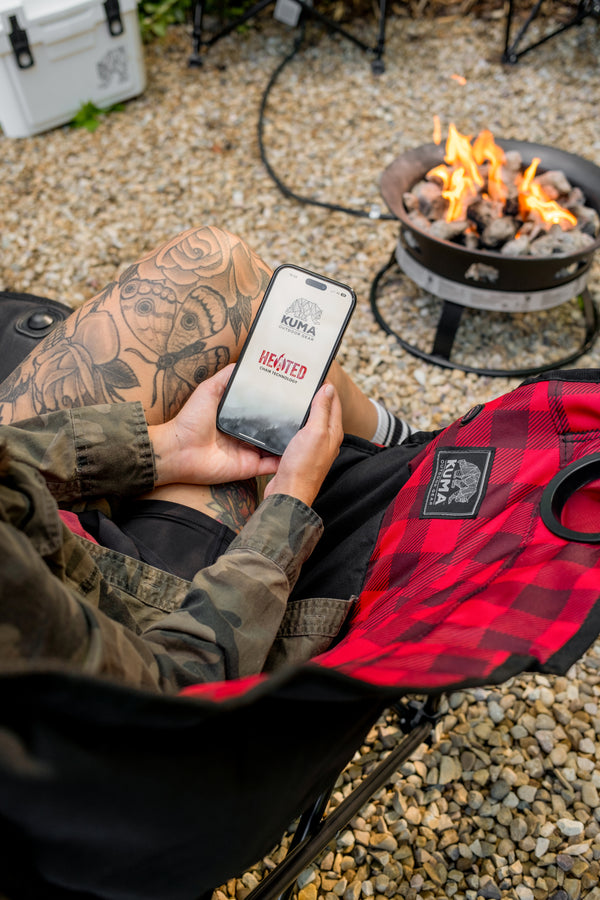 A person sitting in a camp chair outdoors near a fire pit, holding a smartphone with a KUMA branded screen. A red and black plaid shirt is draped over the lap, and the background shows gravel, a cooler, and a burning fire pit.