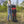 A woman in a gray sweatshirt and blue jeans stands in a grassy outdoor field with trees and mountains in the background, holding a black and red buffalo plaid folding stroller.