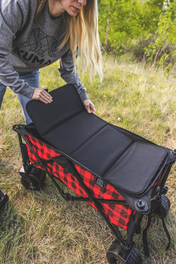A woman outdoors in a grassy field is demonstrating a black padded camping mat laid over a red and black buffalo plaid collapsible wheeled storage cart.