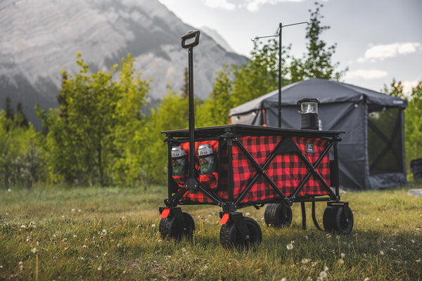 A red and black buffalo plaid folding camping wagon with a black frame and wheels, parked in a grassy field with mountains and a gray tent in the background.