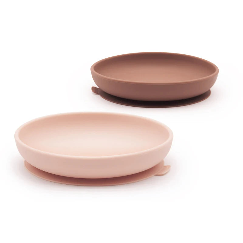 Two round silicone suction plates from the Ekobo Silicone Suction Plate Set for Kids, one in light pink and the other in brown, each featuring raised edges and small side tabs, are shown on a white background. Made from food-grade silicone, this set is designed to provide safe and convenient mealtimes for toddlers.