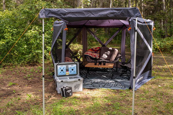 A dark gray mesh camping shelter with a roof and support poles is set up in a wooded outdoor area. Inside the shelter, camping chairs, a small table, and a blanket are arranged.