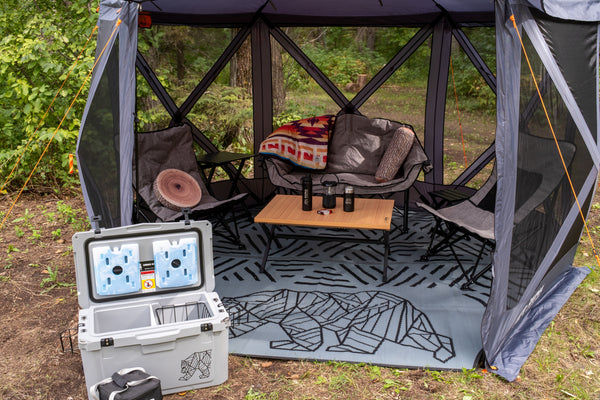A gray outdoor mesh canopy tent with a geometric patterned floor mat, set up in a wooded area. Inside the tent are two folding chairs, a small wooden table with two black canisters, and a blanket draped over the back.