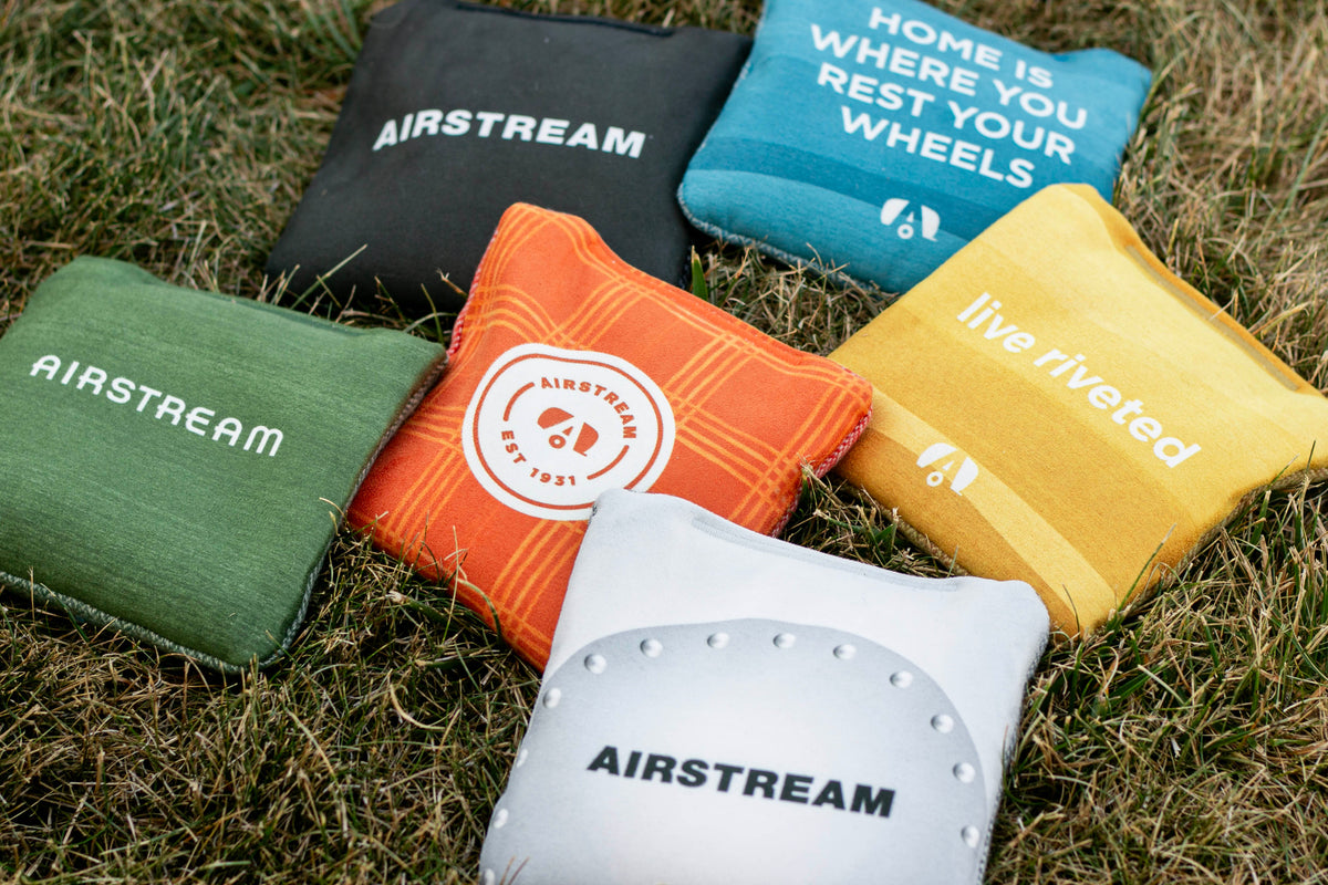 Six colorful Airstream ACA Cornhole Bags—designed as custom bean bags in green, black, orange, blue, yellow, and silver—rest on the grass. Each bag features distinctive Airstream logos and slogans.