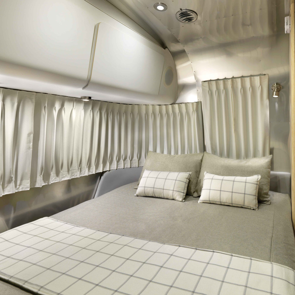 A neatly made bed with light gray bedding and two plaid pillows sits inside a modern, compact camper, featuring Airstream Solid Custom Curtains for Globetrotter Travel Trailers and overhead storage, with the stylish window treatments adding a touch of elegance to the cozy space.