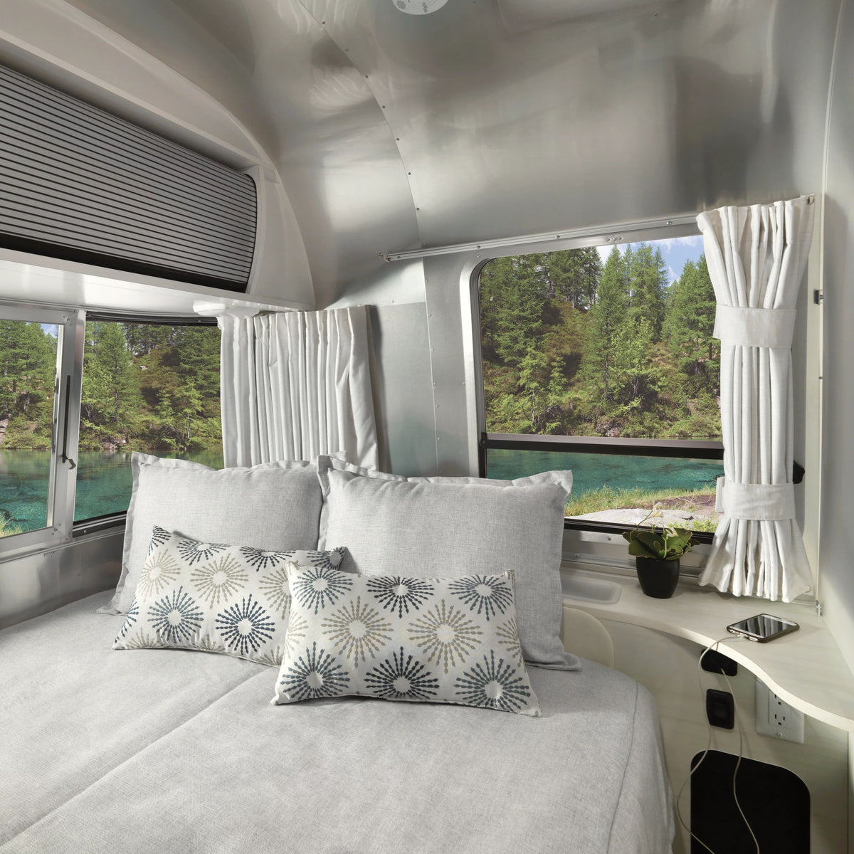 A cozy bed with patterned pillows sits inside a camper van, where large windows fitted with Airstream Solid Custom Curtains for Bambi Travel Trailers frame a scenic view of a lake and pine trees. A small table nearby holds a plant and a smartphone.