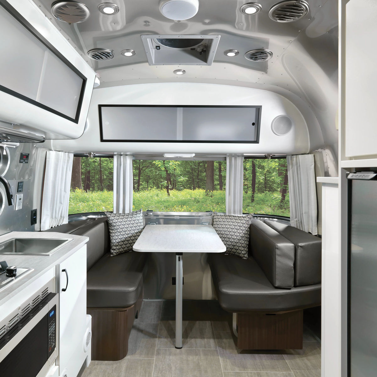 This modern RV interior features a small kitchen on the left and a dining table with two cushioned bench seats, accented by large windows dressed in Airstream Solid Custom Curtains for Caravel Travel Trailers, with green trees visible outside. The space appears clean, bright, and compact.