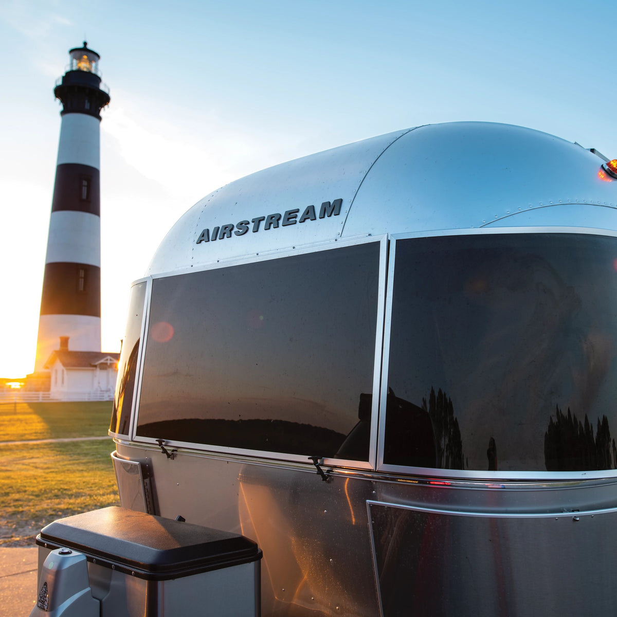 A gleaming Airstream trailer adorned with bold Airstream Logo Decal Black Chrome (Part #386272, 386205-01) is parked near a black-and-white striped lighthouse at sunset. The sky is clear, and sunlight shimmers across the trailer’s metallic finish.