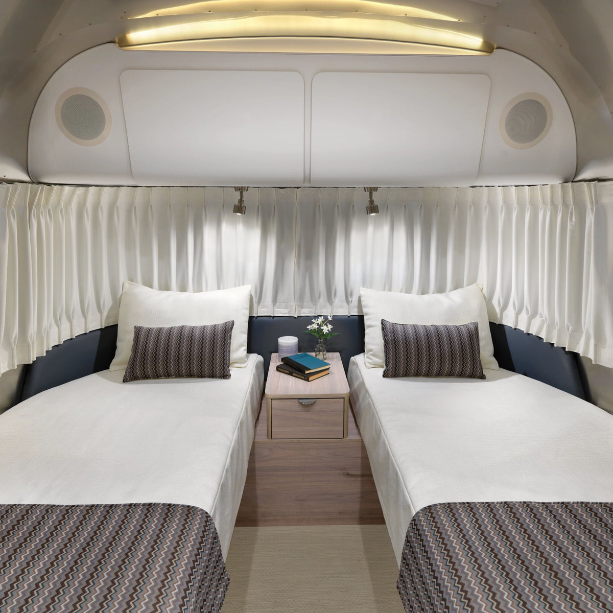 Two twin beds with white linens and pillows are separated by a nightstand holding books, a plant, and a lamp inside a camper van featuring closed Airstream Solid Custom Curtains for Globetrotter Travel Trailers and overhead lighting.