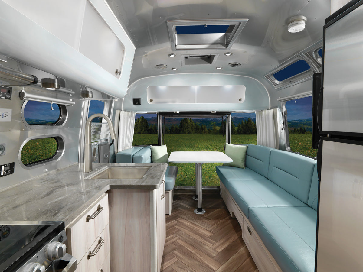 Modern RV interior featuring a kitchen with a sink, marble-style countertops, turquoise seating, and a dining table equipped with the Airstream Threaded and Lock Table Leg System (Part #382722-02, 382723, 382724). Large windows showcase scenic views of grassy fields, trees, and distant mountains beneath a blue sky.