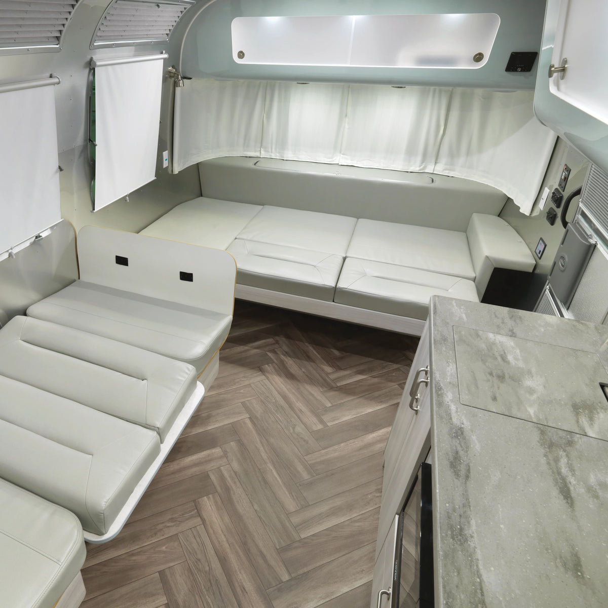 Modern RV interior featuring two convertible cream sofas, a wood-patterned floor, grey counters, and white walls with windows covered by Airstream Solid Custom Curtains for International Travel Trailers. The layout is compact and efficient for both living and sleeping spaces.