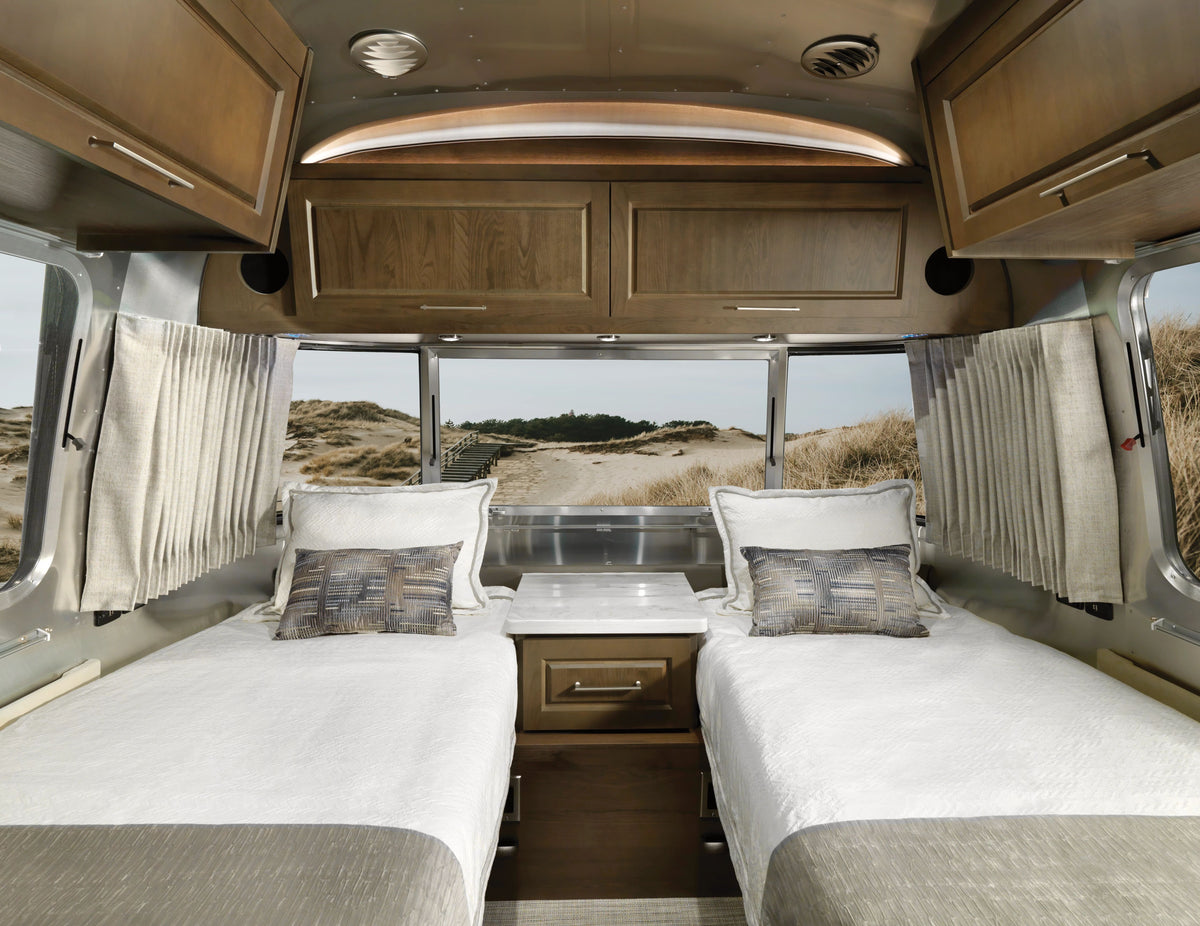 Two twin beds with white bedding inside an RV, featuring wooden cabinets above and windows adorned with Airstream Solid Custom Curtains for Classic Travel Trailers, revealing a view of the sandy beach and grassy dunes outside.
