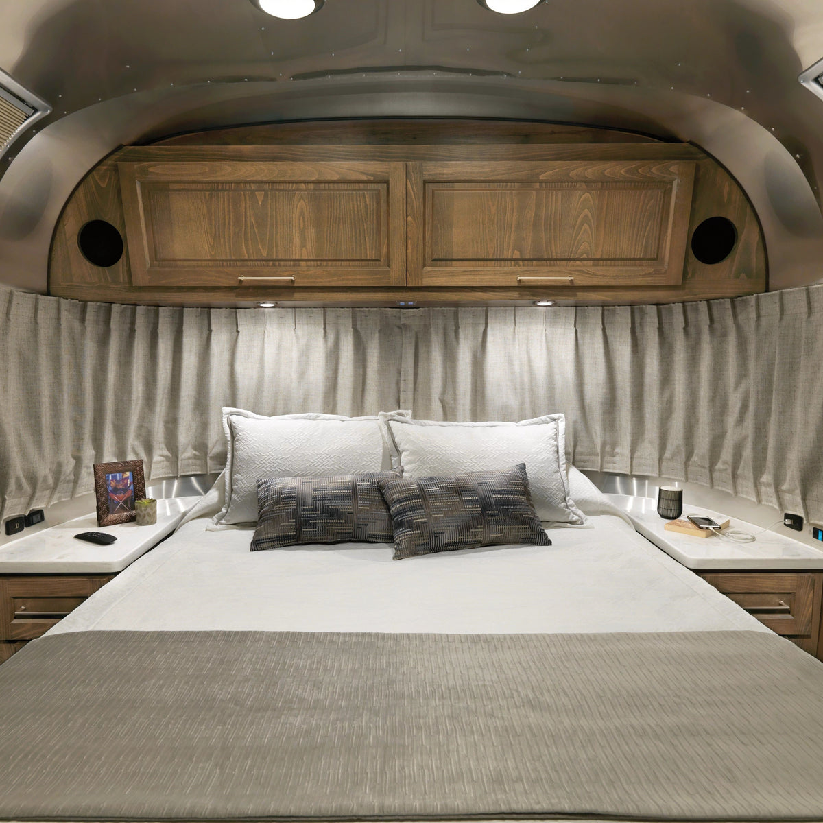A cozy bed with gray pillows and bedding sits beneath wooden cabinets in a modern RV, while Airstream Solid Custom Curtains for Classic Travel Trailers cover the window behind the bed, and small items rest on nightstands on either side.