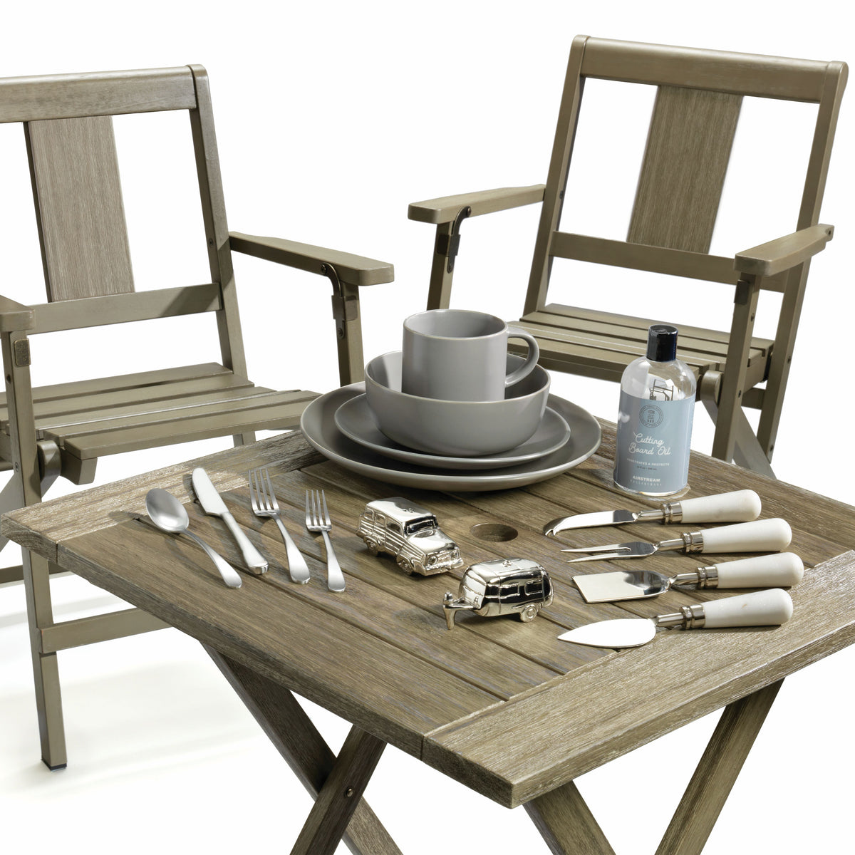 Two Airstream x Pottery Barn Outdoor Chairs flank a wooden table set with grey dishes, a bottle of cutting board oil, metal car and camper figurines, various cutlery, and a set of cheese knives with white handles, all set against a white background.