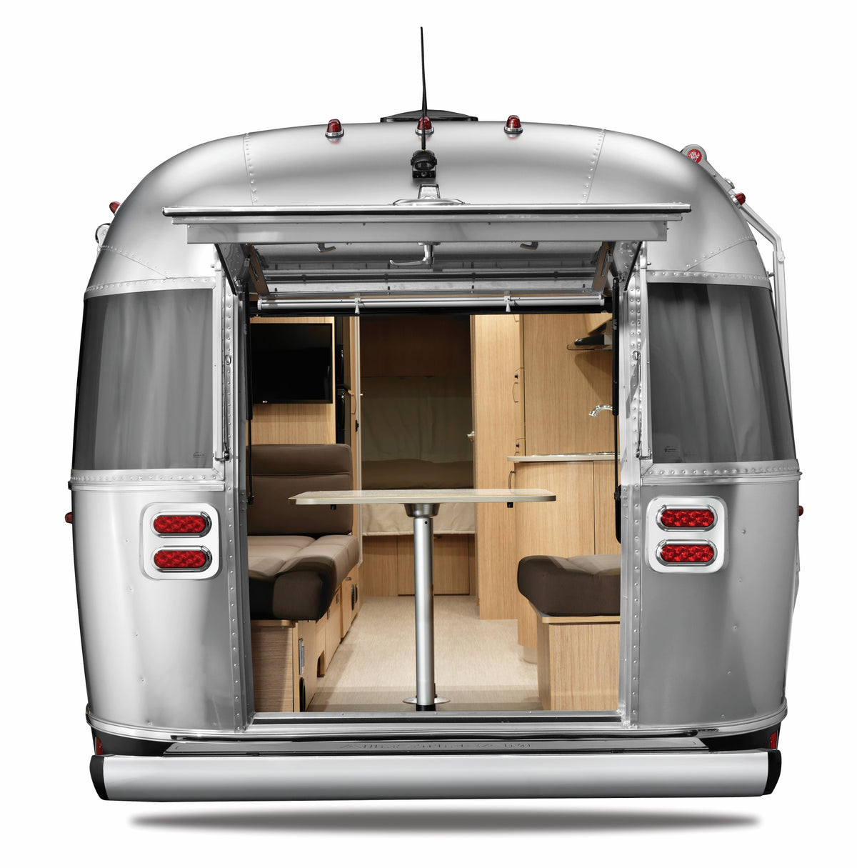 A silver camper trailer with its rear door open, revealing a modern interior featuring brown seating, a central table supported by the Airstream Threaded and Lock Table Leg System (Part #382722-02, 382723, 382724), wood cabinets, and a cozy minimalist design—perfect for those seeking an Airstream interior upgrade.