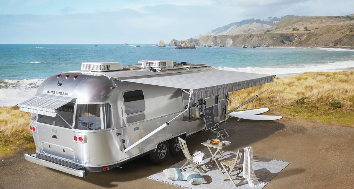 A silver Airstream trailer with an awning is parked by a beach. Next to the trailer, a picnic setup includes the Airstream x Pottery Barn Outdoor Chair Set and a table arranged on a mat beside some surfboards, with ocean waves and cliffs visible in the background.