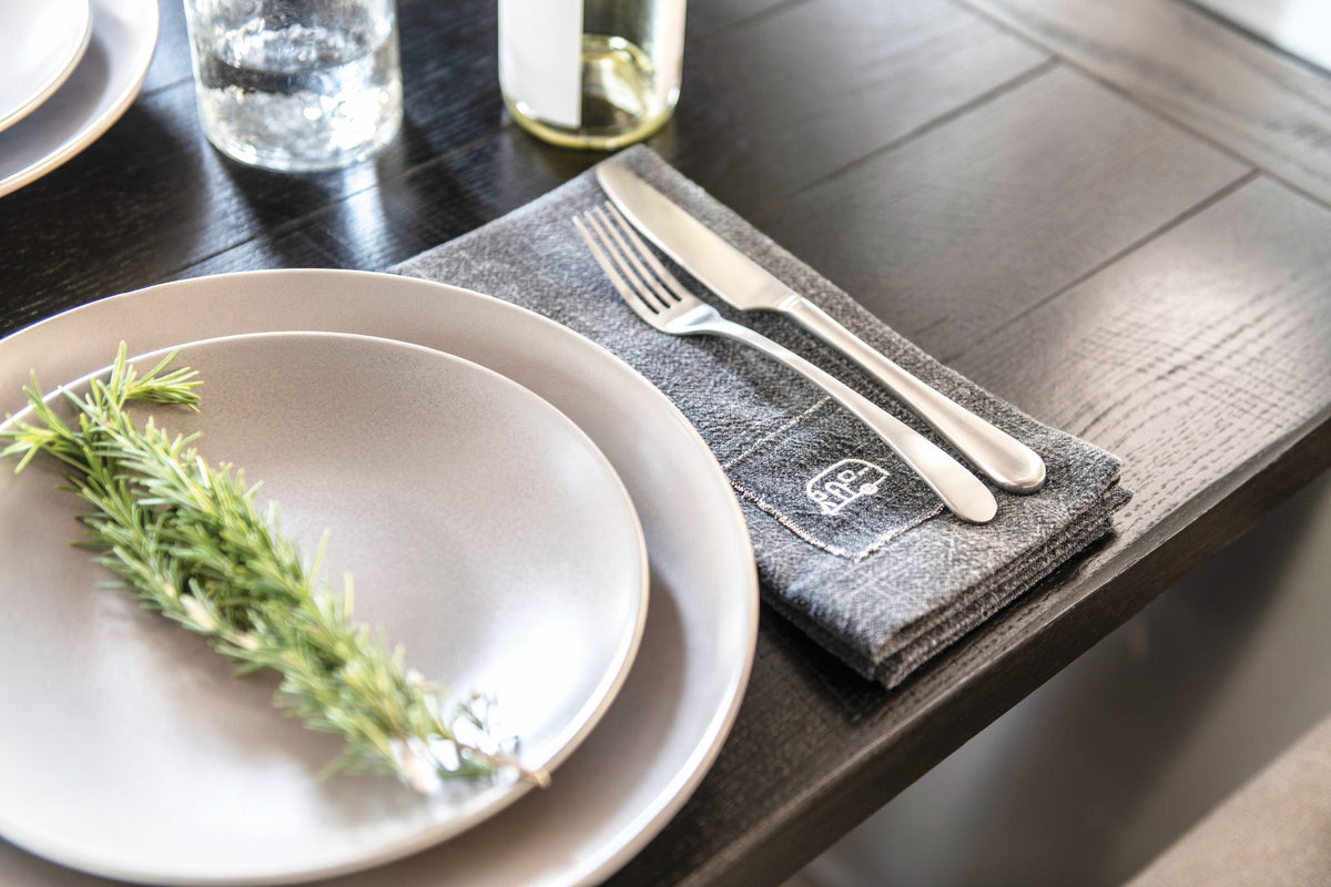 A close-up of a table setting featuring two gray plates from the Airstream x Pottery Barn Dinnerware 16 Pc Set, paired with a sprig of rosemary, a gray napkin with logo detailing, and a fork and knife on a dark wooden table. In the background, a glass and bottle add to the elegant Mason stoneware-inspired aesthetic.
