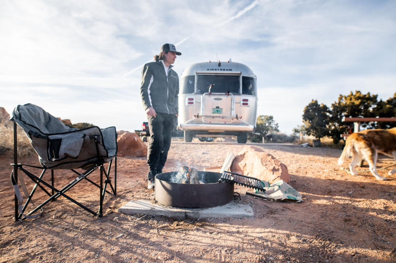 A man stands at a campfire in a desert setting with an Airstream trailer, a folding camp chair, and a dog in the background.