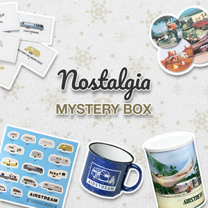 Nostalgia Mystery Box image with vintage travel stickers, postcard, Airstream mug, trailer decals, and a cup featuring a hammock scene, arranged on a snowy background.