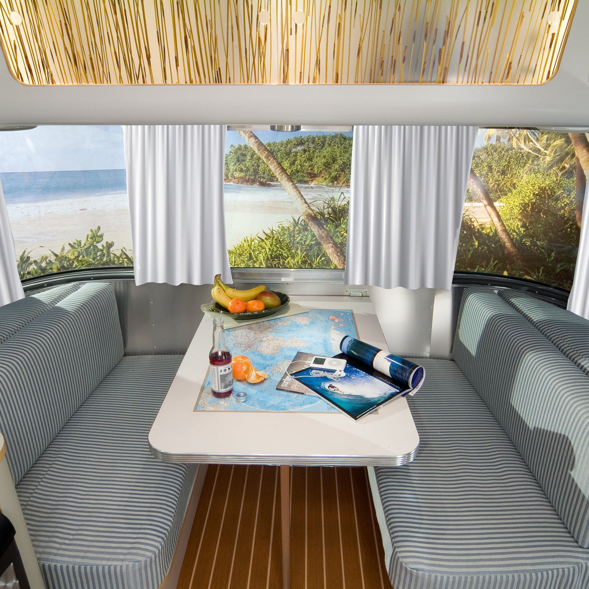 A cozy RV dining area with striped seats and a table set with fruit, a map, a drink, and magazines. Expansive windows are adorned with Airstream Solid Custom Curtains for Quiksilver Travel Trailers, framing breathtaking views of a tropical beach with palm trees and the ocean—an ideal spot to relax in this Quiksilver Special Edition retreat.