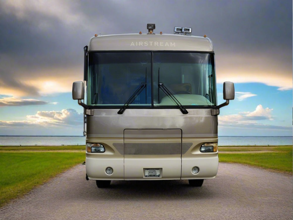A beige Airstream motorhome is parked on a paved road flanked by green grass, with a body of water and cloudy sky in the background—ideal for relaxing inside on its Airstream Custom Mattress for A-Series Motorhomes.