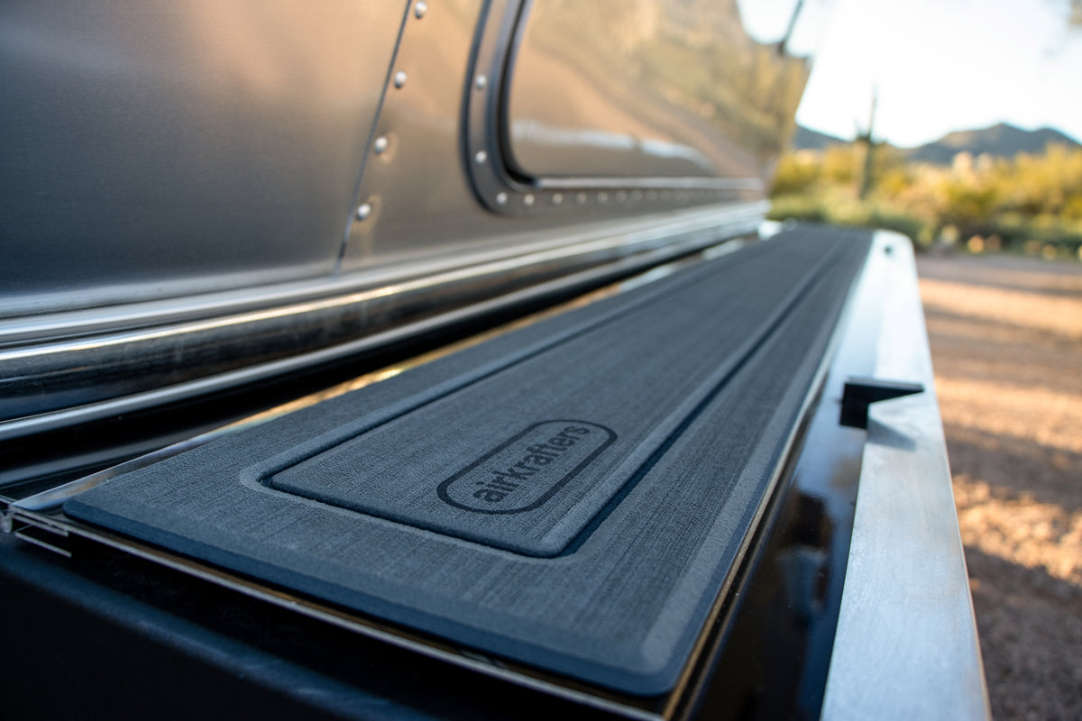 A close-up of the textured bumpMAT™ by Airkrafters, designed as a protective mat for travel trailer accessories, is placed on a metallic surface with a blurred natural background and a shiny metal wall—perfect for use as an Airstream bumper mat.